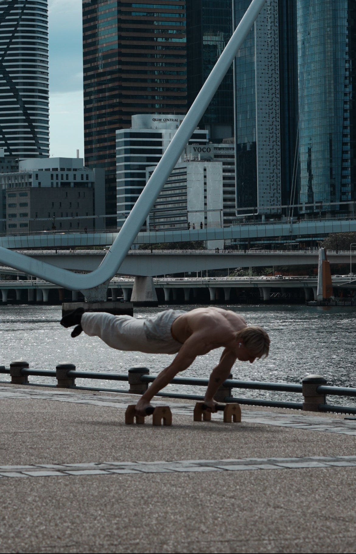 A shirtless man performing a full planche on wooden parallettes along a waterfront, with a modern city skyline and bridge in the background.